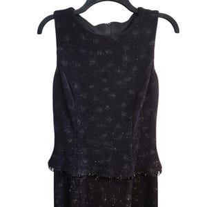 Könnte beinhalten: Schwarzes ärmelloses Kleid mit dezentem Glitzereffekt. Das Kleid hat ein figurbetontes Oberteil mit Schößchen und eine Perlenborte an der Taille. Es hängt an einem schwarzen Kleiderbügel.