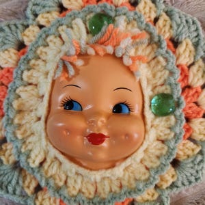 May include: A vintage doll face with blue eyes and red lips, surrounded by a crocheted flower design in pastel colors. The flower has green, yellow, and orange yarn, with two green glass embellishments. The doll has a cheerful expression.
