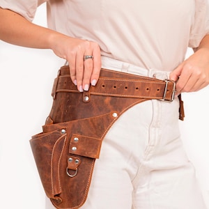May include: A brown leather tool belt with a large pocket on the side. The belt is worn around the waist and has a buckle closure.
