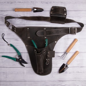 May include: A brown leather gardening tool belt with a variety of pockets and loops for holding tools. The belt is shown with a pair of green pruning shears, a small trowel, a hand rake, and a small hand cultivator.