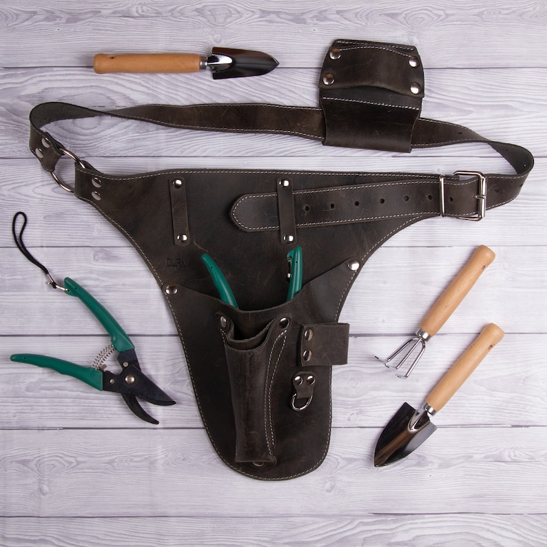 May include: A brown leather gardening tool belt with a variety of pockets and loops for holding tools. The belt is shown with a pair of green pruning shears, a small trowel, a hand rake, and a small hand cultivator.