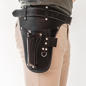 May include: A brown leather tool belt with a stitched design and a metal buckle. The belt has a large pouch with a flap and a smaller pouch with a metal ring.