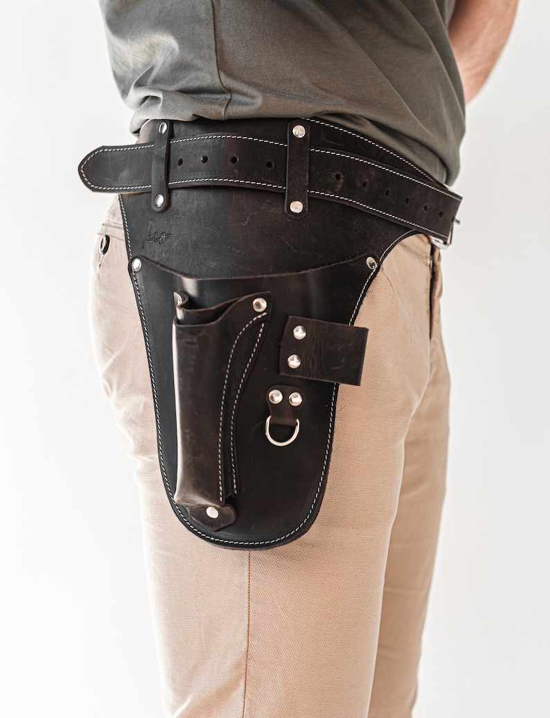 May include: A brown leather tool belt with a stitched design and a metal buckle. The belt has a large pouch with a flap and a smaller pouch with a metal ring.