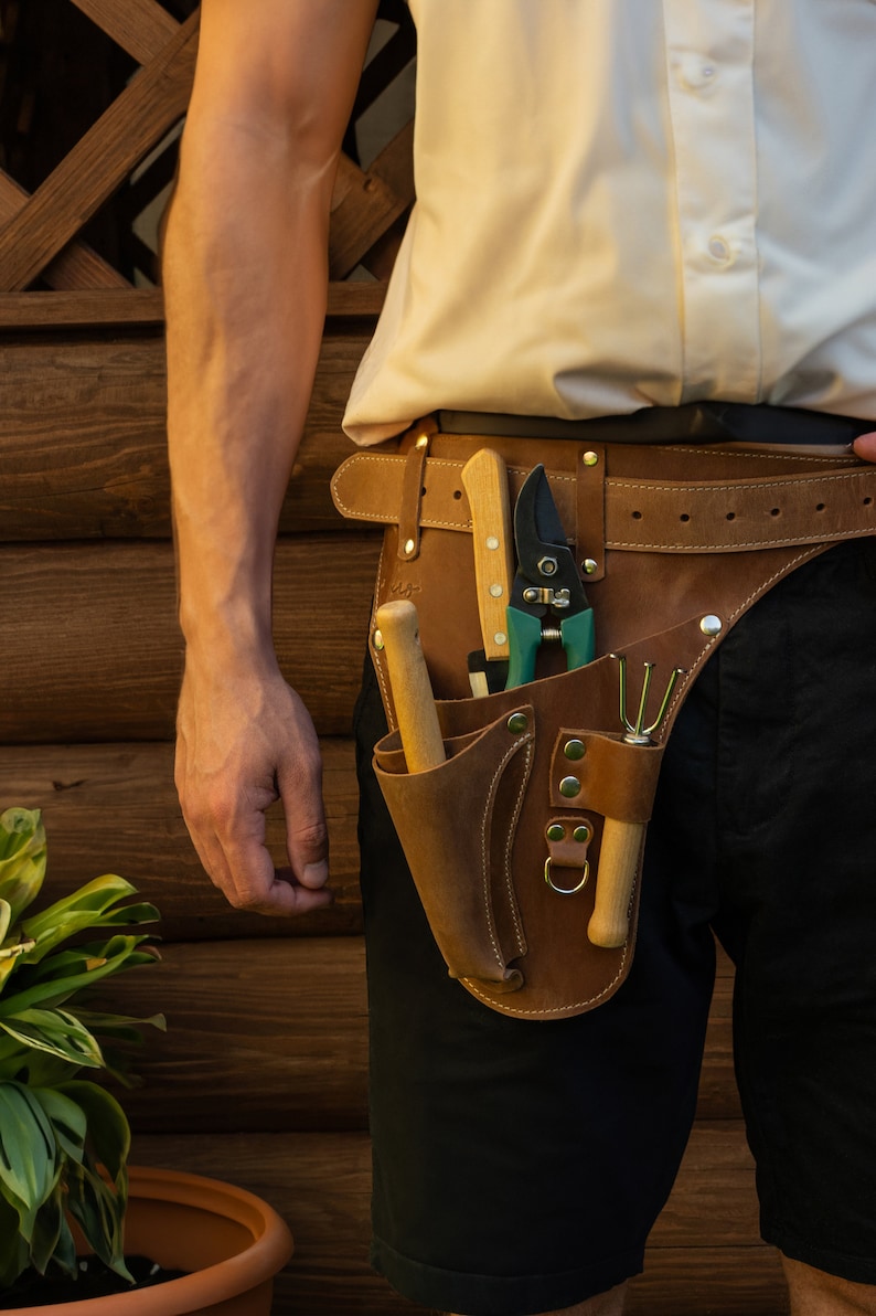 May include: A brown leather tool belt with a variety of gardening tools, including a pair of pruning shears, a small trowel, and a wooden handle.