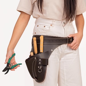 May include: A brown leather gardening tool belt with a brown leather pouch and three gardening tools. The belt is worn around the waist of a person wearing white trousers.
