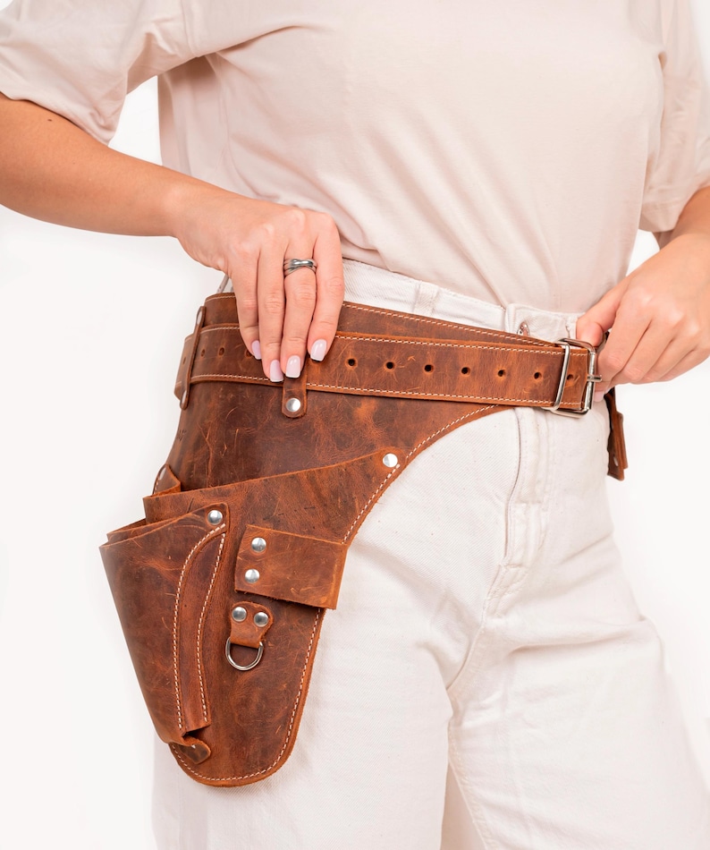 May include: A brown leather tool belt with a large pocket on the side. The belt is worn around the waist and has a buckle closure.