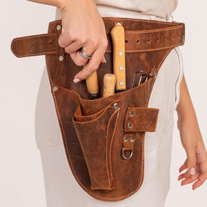 Peut inclure: Une ceinture à outils en cuir marron avec une grande poche et plusieurs petites poches. La ceinture est portée autour de la taille et possède une boucle en métal. La ceinture contient divers outils de jardinage, notamment une paire de sécateurs, une truelle et un râteau à main.