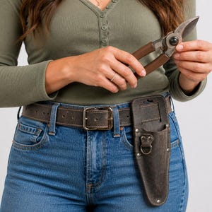 May include: A pair of gardening shears with brown handles and a leather holster attached to a brown belt. The belt has a large square buckle and is worn with blue jeans and a green long-sleeve shirt.