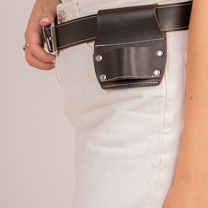 May include: A brown leather belt with a small brown leather pouch attached. The pouch has a flap with a snap closure and is secured to the belt with two metal rivets. The belt is worn over a pair of white jeans.