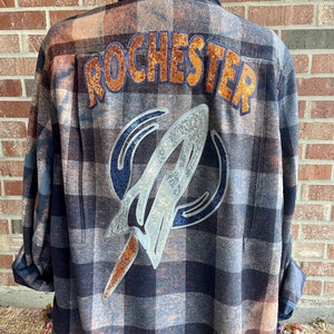 May include: A blue and orange plaid flannel shirt with the word "ROCHESTER" in orange glitter on the back. A white and silver rocket ship design is also on the back.