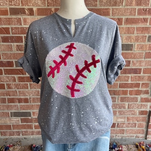 May include: A gray t-shirt with a sparkly white baseball with red stitching on the front. The shirt has a V-neck and short sleeves.