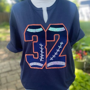 May include: A navy blue short-sleeved t-shirt with a large sequined number "32" in orange and white. The number has a baseball stitch design.