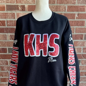 May include: Black sweatshirt with red glitter lettering that says "KHS" and "Mom" on the front. The sleeves have red glitter lettering that says "Lady Tigers 22" on the left sleeve and "Emory Tigers Cheer" on the right sleeve.