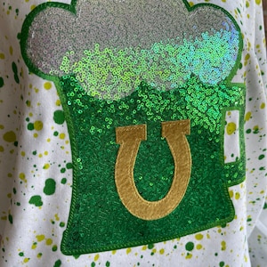 May include: A white shirt with green and yellow paint splatters. A green and gold sequin patch of a beer mug with a shamrock on top is sewn on the front of the shirt.