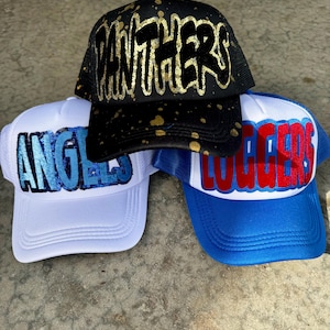 May include: Three trucker hats with mesh backs. One black hat with gold "PANTHERS" lettering and gold splatter. A white hat with blue "ANGELS" lettering. A blue and white hat with red "LOGGERS" lettering.
