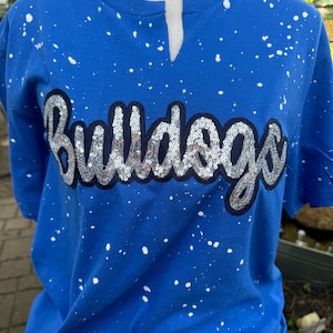 May include: A blue t-shirt with white paint splatters and the word "Bulldogs" in silver sequins. The shirt has a v-neck design.