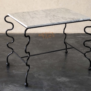 Op de afbeelding: Een rechthoekige bijzettafel met een wit marmeren blad met grijze aders. Het onderstel is gemaakt van zwarte metalen buizen in een doorlopend, verticaal golvend patroon aan elke kant, rustend op vier kleine bolvormige voetjes aan de basis.
