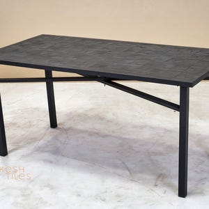 Puede incluir: Una mesa rectangular negra con una superficie texturizada y patas de metal negro. La mesa mide aproximadamente 152 cm de largo y 76 cm de ancho. La mesa está en una habitación con suelo claro y paredes de tonos neutros. El texto "kesh tiles" es visible.