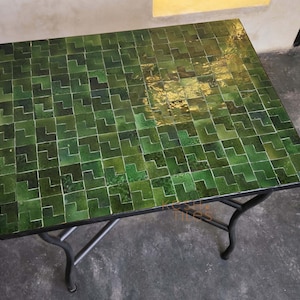 Puede incluir: Una mesa cuadrada con una tapa de mosaico de azulejos verdes. Los azulejos están dispuestos en un patrón repetitivo. La mesa tiene una estructura y patas de metal negro. La mesa mide aproximadamente 76 cm de largo y 76 cm de ancho.