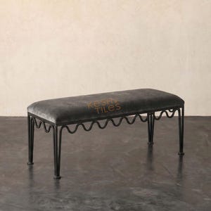 May include: A rectangular bench featuring a dark gray upholstered seat and a black metal frame with a distinctive wavy, scalloped design along the sides. The text KESHI TILES is printed in a light brown color on the center of the seat cushion.
