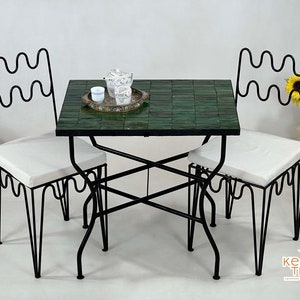 Pode incluir: Uma mesa de metal preto com tampo de azulejos verdes e duas cadeiras combinando. As cadeiras t&ecirc;m almofadas brancas e um design ondulado no encosto.