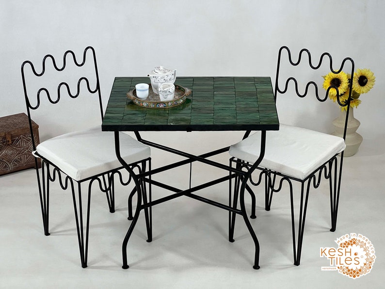 Pode incluir: Uma mesa de metal preto com tampo de azulejos verdes e duas cadeiras combinando. As cadeiras t&ecirc;m almofadas brancas e um design ondulado no encosto.
