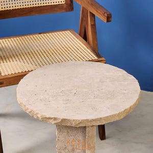 May include: A round, light beige travertine side table with a rough, textured surface. The table has a unique, modern design with a single, thick, rectangular leg.