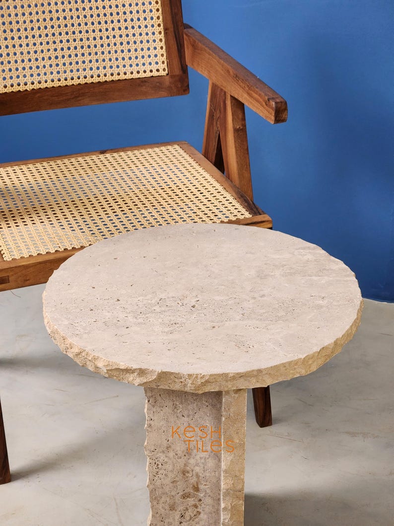 May include: A round, light beige travertine side table with a rough, textured surface. The table has a unique, modern design with a single, thick, rectangular leg.