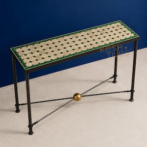 Può includere: Un tavolo console rettangolare con un piano in piastrelle a mosaico con un motivo a rombi color crema e verde. Il tavolo ha una struttura in metallo scuro con quattro gambe dritte e un supporto a croce con una sfera dorata. Testo KESH TILES.