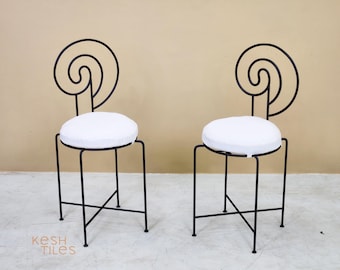Op de afbeelding: Twee zwarte metalen barkrukken met witte ronde kussens. De rugleuningen van de krukken hebben een decoratief wervelontwerp. De krukken hebben een minimalistisch design en staan tegen een neutrale achtergrond.