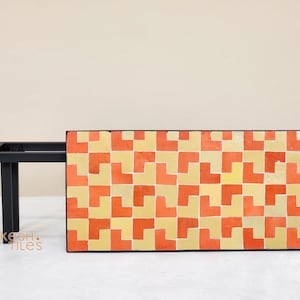 May include: A rectangular coffee table with a black metal frame and a patterned top. The top features a repeating geometric design in shades of orange and yellow. The table is a modern design.