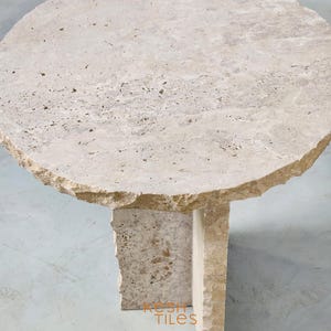 May include: A round, light beige travertine side table with an irregular, rough-hewn edge. The table has a simple, modern design with a single, rectangular base.