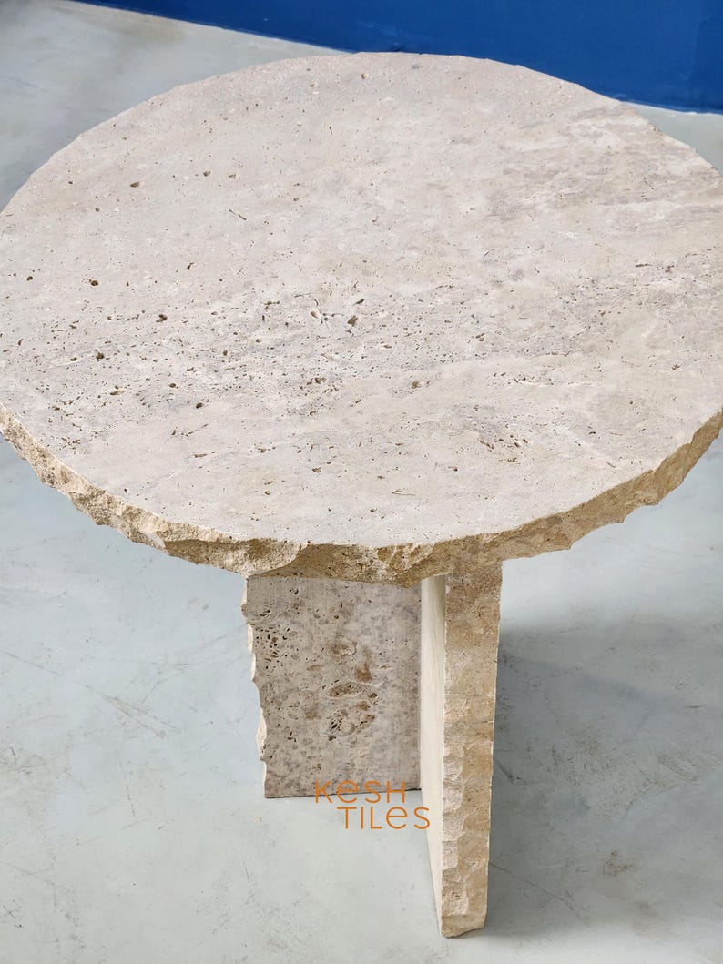 May include: A round, light beige travertine side table with an irregular, rough-hewn edge. The table has a simple, modern design with a single, rectangular base.