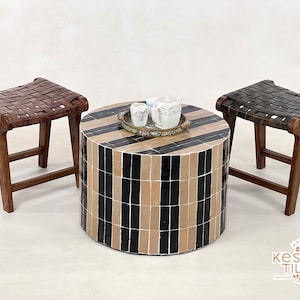 May include: Three wooden stools with woven leather seats and a round black and beige tiled coffee table with a decorative tray and teacups on top. The table and stools are arranged in a symmetrical pattern on a white surface.