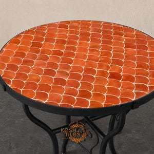 Può includere: Un tavolo rotondo con piano rivestito da piastrelle arancioni a forma di squama di pesce con fughe bianche. Il tavolo ha una struttura in metallo nero con gambe curve. Un logo con il testo KESH TILES è visibile sul telaio inferiore.