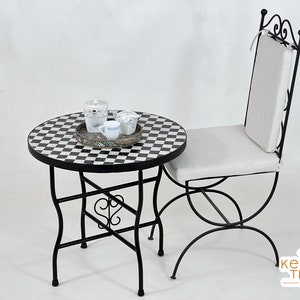 Puede incluir: Una mesa de bistro de metal negro con una tapa de azulejos en blanco y negro a cuadros. La mesa tiene un diseño decorativo de desplazamiento en las patas. Una silla acolchada blanca con un marco de metal negro se encuentra junto a la mesa.