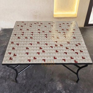 Peut inclure: Une table carrée dotée d'un plateau en mosaïque avec un motif répétitif de carreaux rectangulaires gris clair et blancs, agrémenté de petits carreaux carrés rouge foncé dispersés. La table repose sur un cadre en métal noir à quatre pieds.