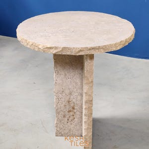 May include: A round, light beige stone side table with a unique, irregular, and textured design. The table has a circular top and a square base with a unique, irregular, and textured design. The table is made of travertine stone.