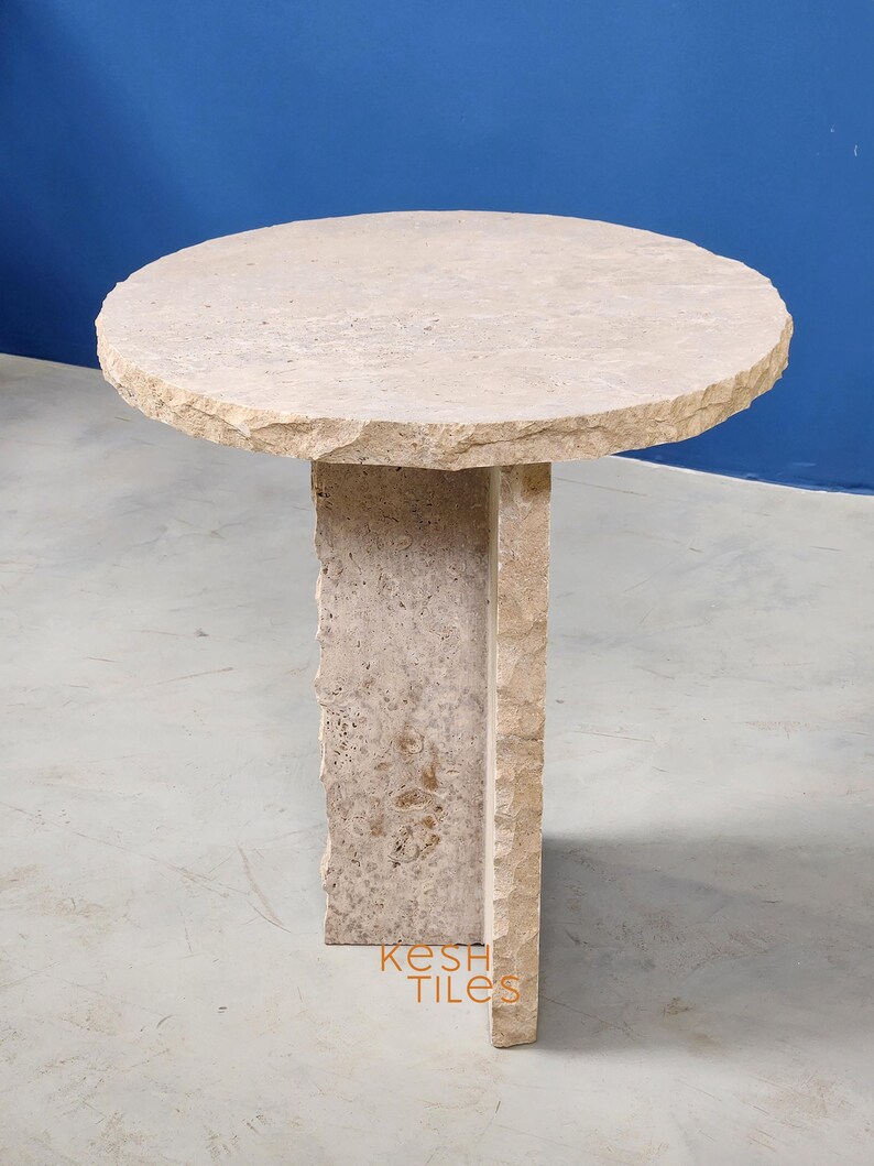 May include: A round, light beige stone side table with a unique, irregular, and textured design. The table has a circular top and a square base with a unique, irregular, and textured design. The table is made of travertine stone.