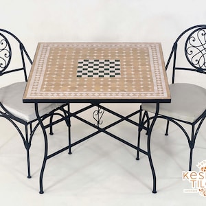 Könnte beinhalten: Ein schwarzes Schmiedeeisen-Terrassenset mit einem quadratischen Tisch und zwei Stühlen. Der Tisch hat eine Mosaik-Tischplatte mit einem Schachbrettmuster in der Mitte und einem beige-weißen Muster. Die Stühle haben graue Kissen und dekorative Rückenlehnen.