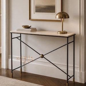 May include: A rectangular console table with a light-colored stone top and a dark metal frame featuring an X-shaped crossbar design with a central sphere. The legs have brass-toned feet. The text KESH TILES is visible on the lower left side.