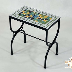 Puede incluir: Una pequeña mesa de metal negro con una tapa cuadrada hecha de azulejos de mosaico verde, amarillo y azul. La mesa tiene un diseño sencillo con dos patas curvas a cada lado.