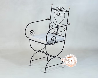 May include: A black metal chair with a white cushion and intricate scrollwork design. The chair has a curved back and arms, and the legs are made of metal.