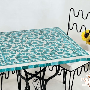 May include: A square table with a teal and white Moroccan tile top and a black wrought iron base. Two chairs with black wrought iron frames and white cushions are positioned around the table.