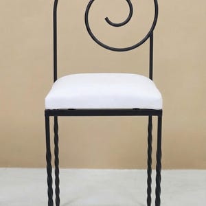 May include: Black wrought iron chair with a white cushion. The chair features a decorative swirl design at the top of the backrest. The legs have a twisted rope design. The chair is suitable for indoor use.