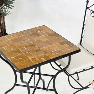 May include: A square table with a black metal frame and a top made of brown mosaic tiles. A black metal chair with a white cushion sits next to the table.