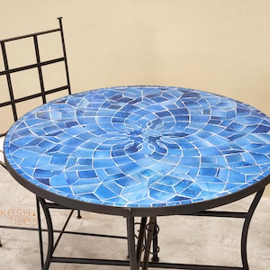 May include: Round outdoor table with a mosaic tile top in shades of blue. The table has a black metal frame and is paired with a matching chair. The mosaic design features a central floral pattern.