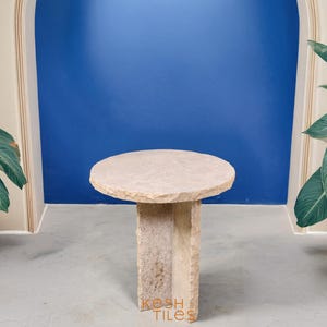 May include: A round, light beige stone side table with a rough, uneven surface and a unique, asymmetrical base. The table is standing on a light gray concrete floor.