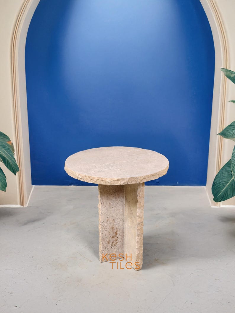 May include: A round, light beige stone side table with a rough, uneven surface and a unique, asymmetrical base. The table is standing on a light gray concrete floor.