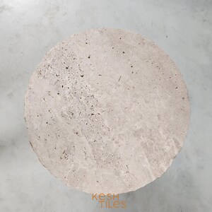 May include: A round, light beige travertine stone table top with a rough, textured surface. The table top is approximately 18 inches in diameter.